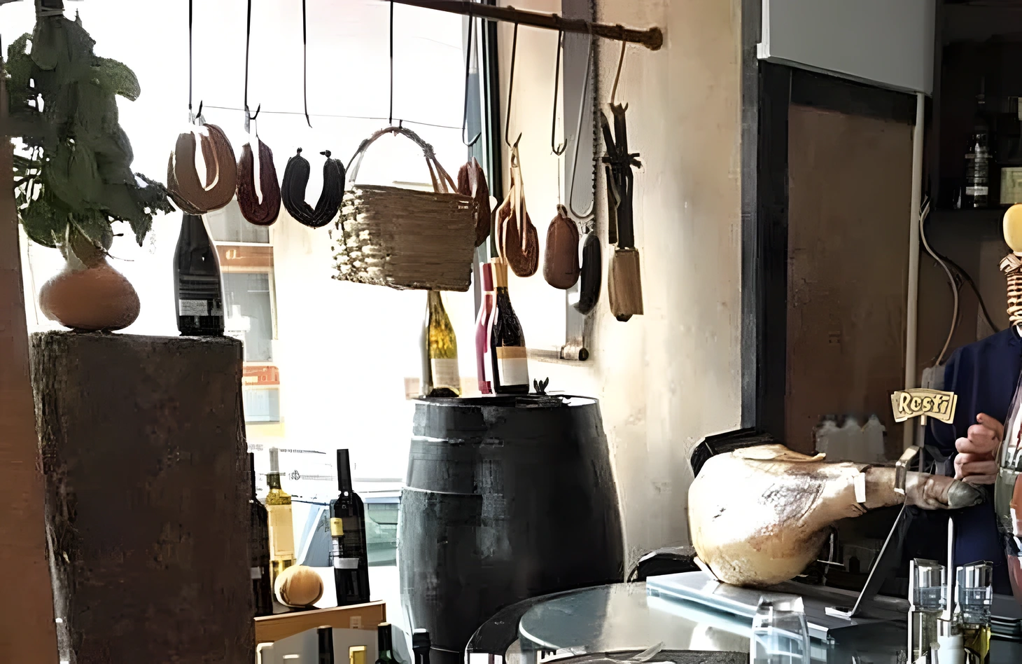Chouriços e linguiças pendurados num varão de madeira, ao fundo um cesto de vime e garrafas de vinho sobre um barril.