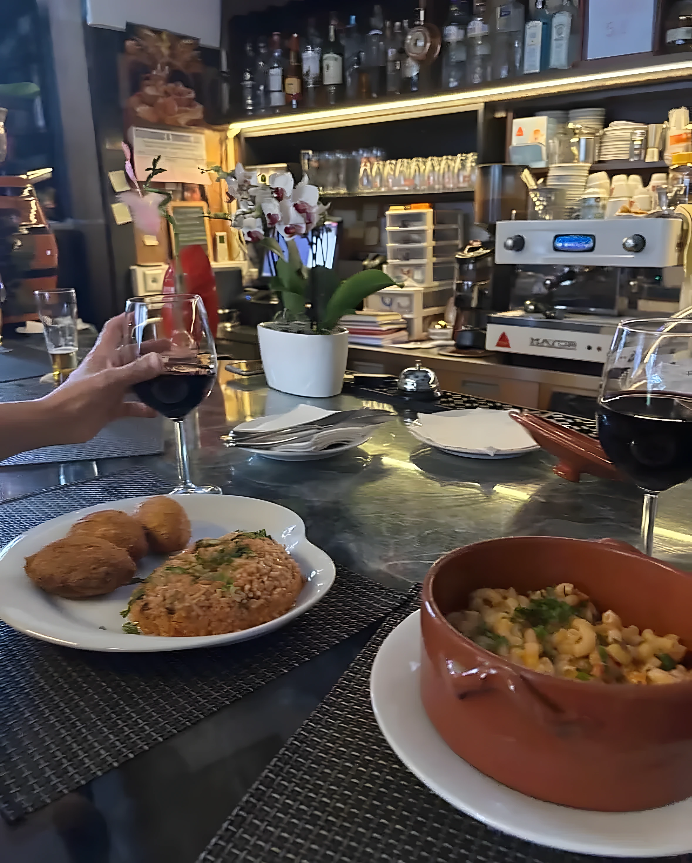 Pastéis de bacalhau com arroz de tomate, uma taça de vinho tinto e uma panela de barro com massa, sobre o balcão de mármore.